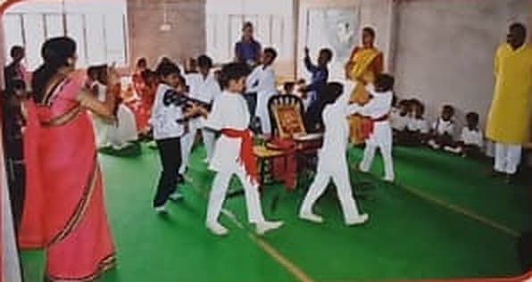 Children in white uniforms with red sashes performing in the school hall while teachers watch.