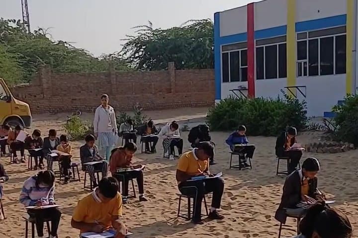 Students writing an examination seated on stools spread across a sandy school ground, in front of a white building with blue, yellow and red stripes.