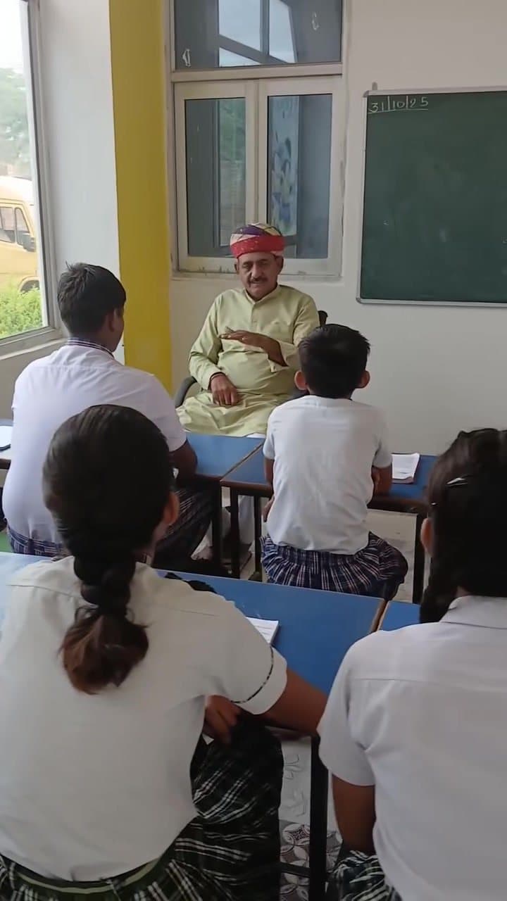 An elder in a red safa talking with students seated at blue desks in a bright classroom.