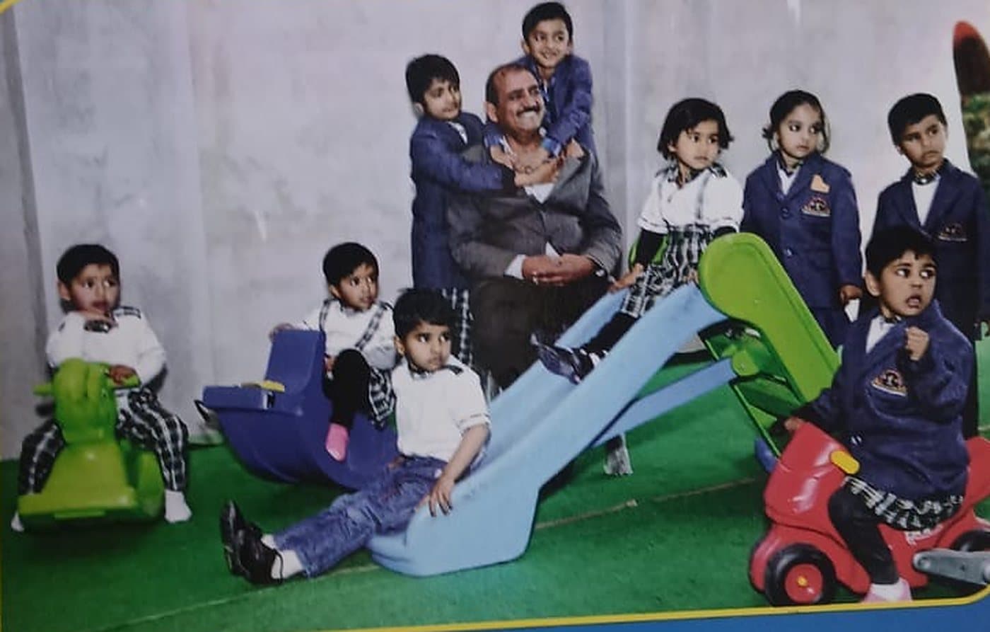 Nursery children in school uniforms playing on a slide and ride-on toys on a green play mat, with a smiling adult seated among them.