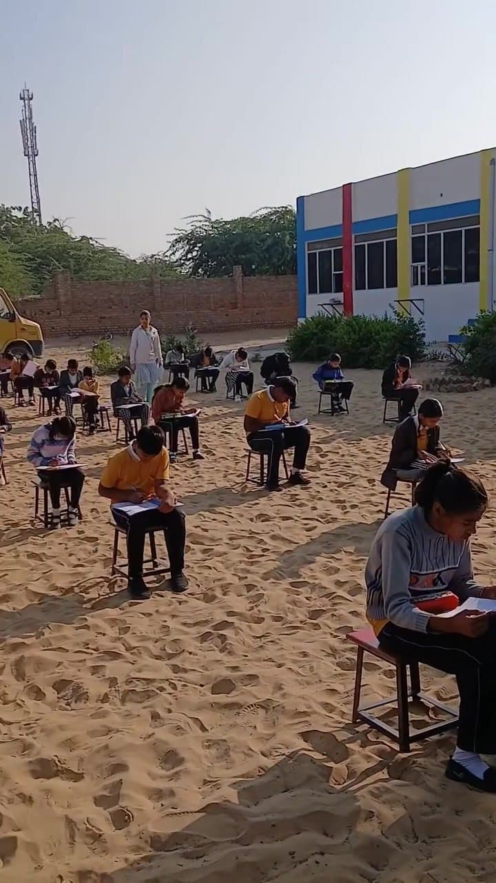 An open-air examination on the sandy school ground with the striped school building behind.
