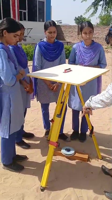 Girls in blue uniforms and dupattas gathered around a plane table on a yellow tripod during a geography practical.