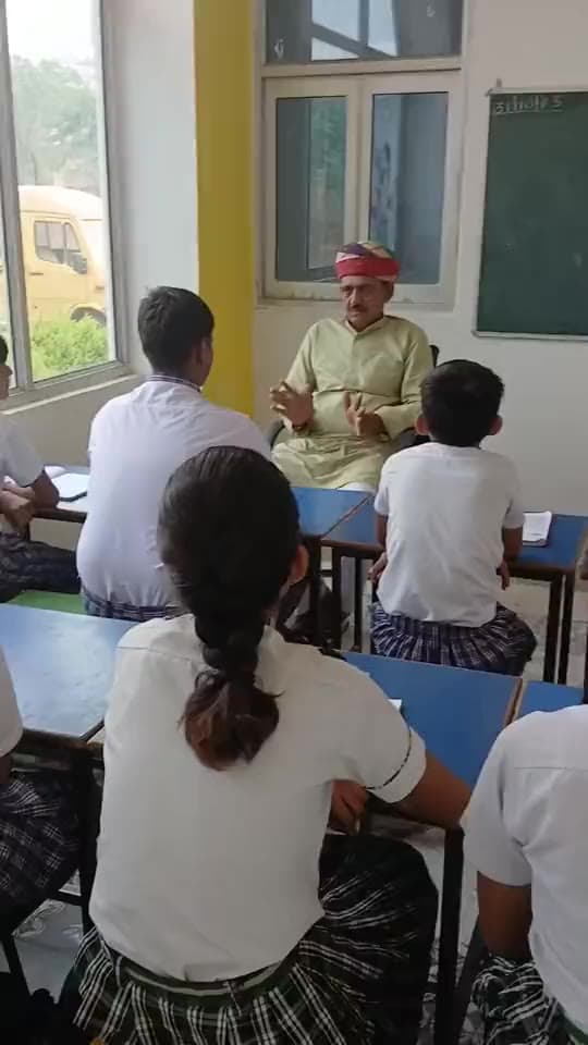 an elder in a red safa speaks with students in a classroom.