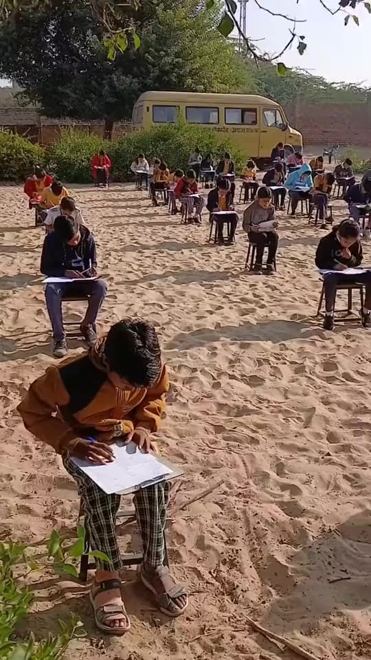 the camera pans across students writing an examination outdoors on the sandy school ground.